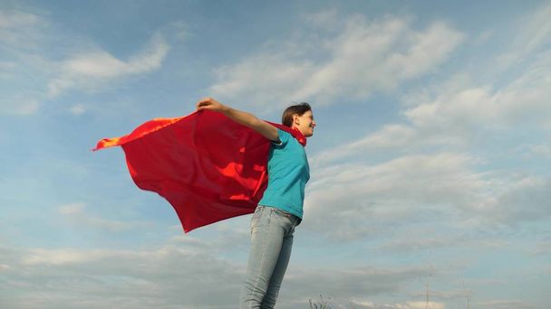 beautiful girl superhero standing on the field in a red cloak, cloak fluttering in the wind. Slow motion. girl dreams of becoming a superhero. young girl standing in a red cloak expression of dreams - Photo, Image