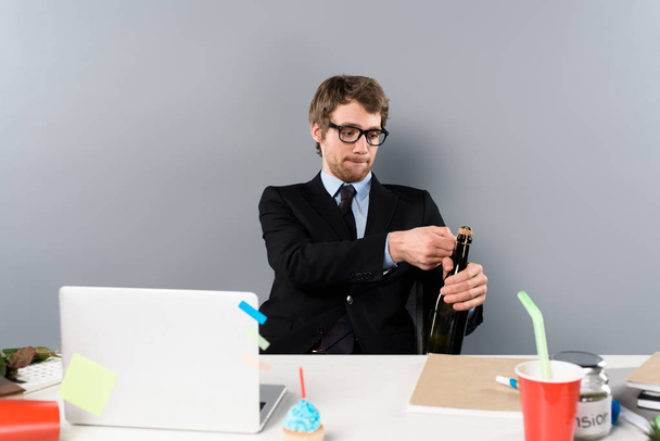 επιχειρηματία που κάθεται στο χώρο εργασίας και άνοιγμα μπουκάλι με σαμπάνια που απομονώνονται σε γκρι - Φωτογραφία, εικόνα