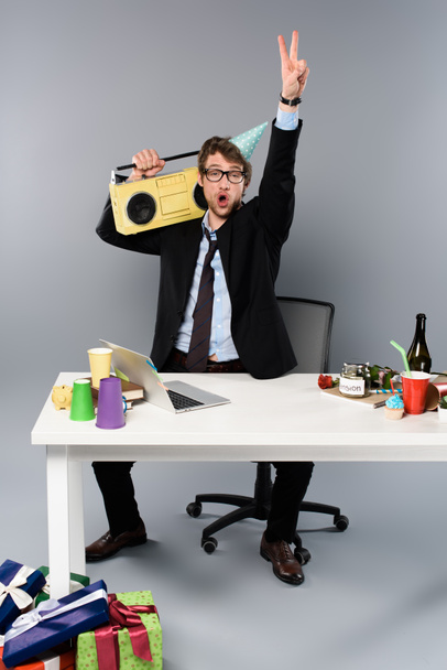 drunk businessman sitting at workplace in party cap with vintage tape recorder on grey background - Photo, Image