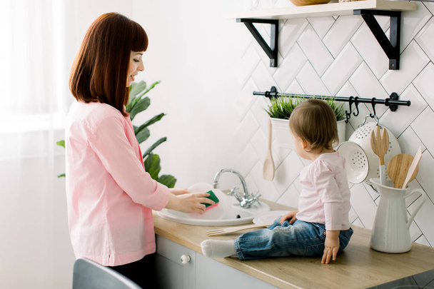 Young happy mother and her cute curly toddler daughter having fun together in a white kitchen. Mother is washing the dishes, plates, while baby girl is sitting near sink and playing with wooden fork - Foto, Imagen