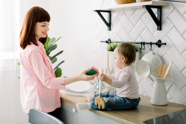 Little baby girl helping her pretty mother washing dishes in the kitchen. Focus on girl. A small child with interest helps her mother with household chores. The girl is happy to learn to wash dishes. - Photo, Image