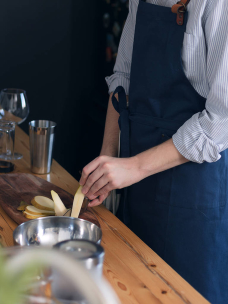 Male bartender in a blue apron in the kitchen, cutting fruits on a wooden cutting board - Fotó, kép