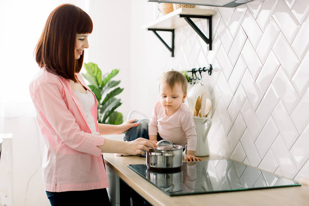 Curious funny kid girl playing with pot with mom in the kitchen, excited active child helping single mother preparing food with cookware, family mommy and daughter having fun cooking together - Photo, image