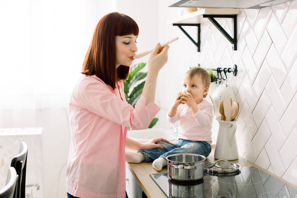 Mother with her daughter in the kitchen cooking together tasty dinner. Little baby girl eats a green apple. Focus on the baby - Fotografie, Obrázek