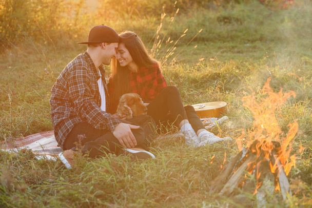 Happy caucasian couple in casual clothes with their funny dog sitting on the lawn in spring park. Young beautiful girl nd her handsome boyfriend leaned against each other at the nature. - Fotó, kép