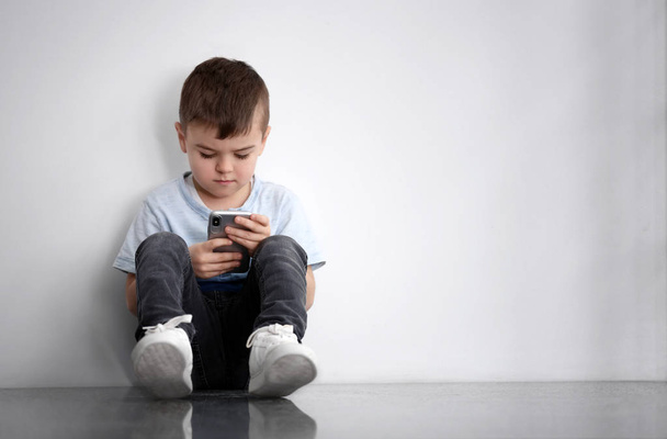 Sad little boy with mobile phone sitting  near white wall, space for text - Zdjęcie, obraz