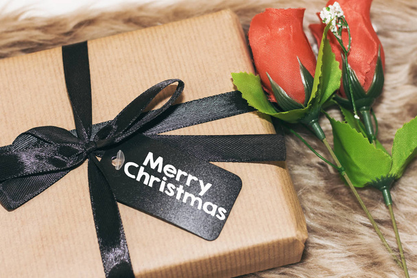 διακοπές έννοια, δώρο κουτί με χαρούμενα Χριστούγεννα tag και κόκκινα ροδαλά λουλούδια πάνω από μια ψεύτικη γούνα - Φωτογραφία, εικόνα
