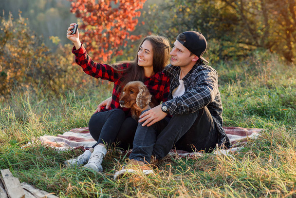 Stylish couple have a video call with their friends. Young happy hipster couple with their dog making selfie using smart phone while sitting on the lawn in spring park. - Fotó, kép