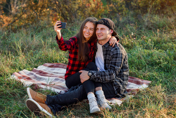 Joven pareja hipster feliz haciendo selfie usando el teléfono inteligente mientras está sentado en el césped en el parque de primavera. Elegante pareja moderna tienen una videollamada con sus amigos sentados en la naturaleza
. - Foto, imagen