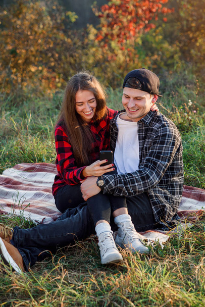 Beautiful girl sits on her boyfriend lap and using smart phone on the lawn in the park. Hipster modern couple have a chatting with their friends sitting in the nature. - Photo, image