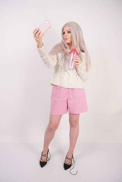 linda menina loira fica na moda roupas casuais, leva uma selfie em seu smartphone e mantém uma xícara transparente rosa no fundo do estúdio branco
 - Foto, Imagem