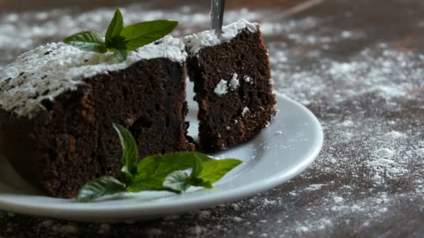 Ciasto domowe pieczone Piernik czekoladowy stłumiony z cukrem pudrem na białym talerzu urządzone z liści mięty. Widelec zrywa kawałek tortu brownie z płytki - Materiał filmowy, wideo