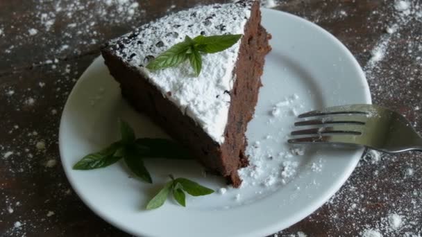 Kawałek czekoladowego ciasta Brownie na białej płycie ozdobione świeżych liści mięty obok widelca - Materiał filmowy, wideo