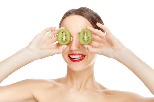 Pretty girl with fresh healthy skin hiding her eyes with kiwi, smiling and showing tip of her tongue - Foto, afbeelding