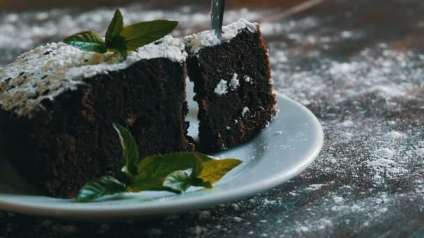 Ciasto domowe pieczone Piernik czekoladowy stłumiony z cukrem pudrem na białym talerzu urządzone z liści mięty. Widelec zrywa kawałek tortu brownie z płytki - Materiał filmowy, wideo