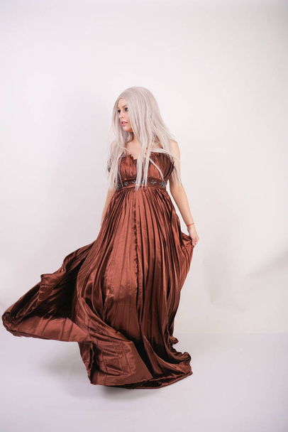 luxury blonde caucasian model girl in chocolate color long evening dress made of pleated fabric waving a flying dress and stands on a white Studio background - Photo, Image