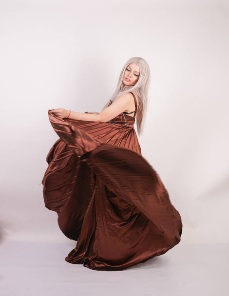 luxury blonde caucasian model girl in chocolate color long evening dress made of pleated fabric waving a flying dress and stands on a white Studio background - Valokuva, kuva