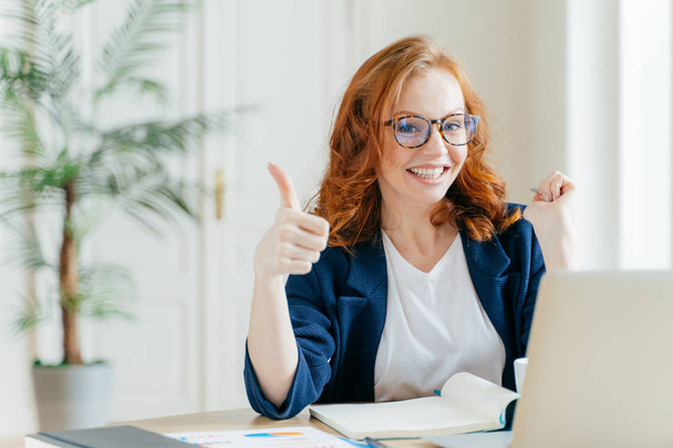 Positive redhead woman keeps thumb raised, demonstrates like gesture, satisfied with good work of colleague, updates software on modern gadget, searches data on website, poses in coworking space - Photo, Image