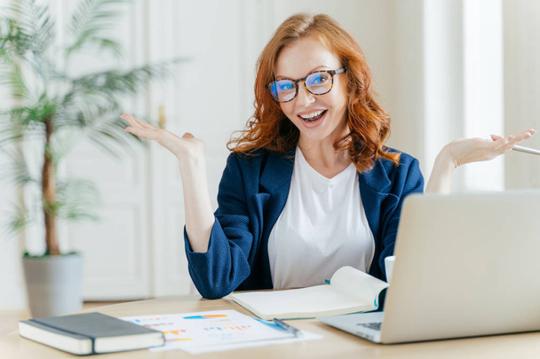 Image of cheerful redhead businesswoman watches webinar or tutorial video, uses free internet connection, raises palms with hesitation, sits at desktop with papers. Female owner of small business - Foto, immagini