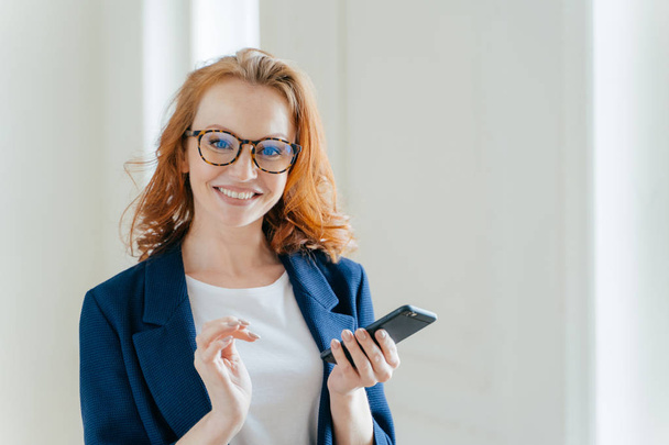 Elegant female copywriter with broad smile searches information for article and publication on cell phone wears spectacles. Businesswoman learns details of business contract online on finance platform - Photo, Image
