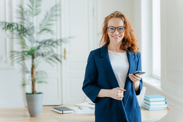 Happy businesswoman reads incoming notification, holds smartphone device, checks email box, has wavy red long hair, dressed in formal clothes, poses over office interior, has confident look at camera - Foto, Imagen