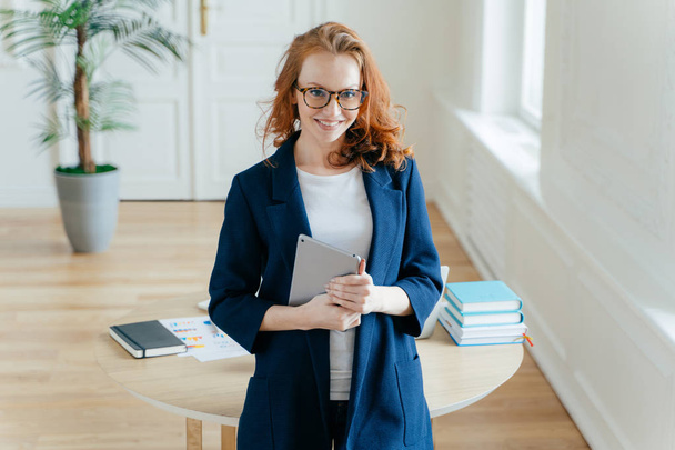 Happy good looking female office worker enjoys online banking service, checks balance on touch pad, reads financial news, wears elegant clothes, stands near table in cozy cabinet, smiles gently - Photo, Image