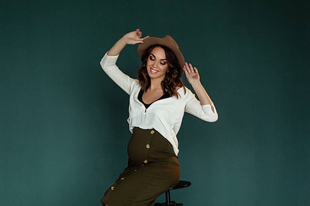 Pregnant european woman in white blouse and green skirt with beige hat in dark flat, stylish and happy pregnant woman in apartments, beautiful pregnant woman in dark studio - Foto, imagen