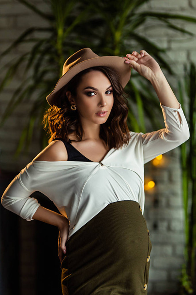 Pregnant european woman in white blouse and green skirt with beige hat in dark flat looks to the camera, stylish and happy pregnant woman in apartments, beautiful pregnant woman in dark studio - Foto, afbeelding