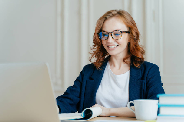 Glad lovely woman has ginger hair, positive smile, sits with laptop computer at desktop, happy to make video call or conference, communicates with investors from abroad, drinks aromatic coffee. - Photo, Image