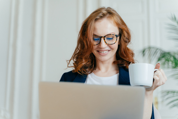 Headshot of smiling red haired female in optical glasses, enjoys hot coffee or tea, focused in laptop computer, makes purchases online, smiles pleasantly, works freelance on distance, books tickets - Photo, Image