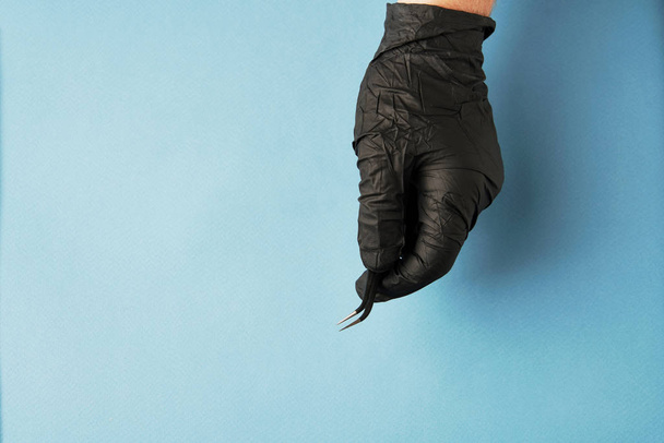 Hand in black glove holding tweezers on light blue background - Photo, Image