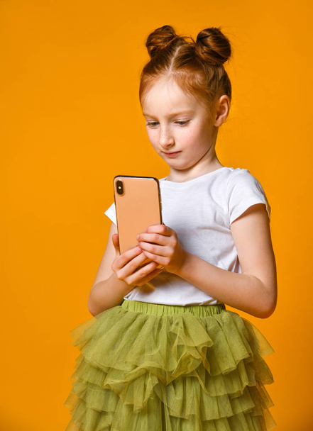 Portrait of a red-haired little girl looking smart phone, standing against a yellow wall - 写真・画像