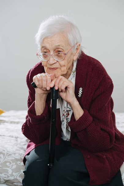 Portrait of a senior woman sitting on bed with walking stick at home - Fotoğraf, Görsel