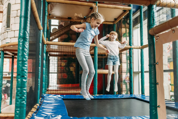 Two funny girls jumping on the trampoline, children game center. Excited childs having fun on playground indoors. Kids playing in amusement centre - 写真・画像