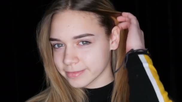 Πορτραίτο ενός όμορφου νεαρού κοριτσιού με μακριά λευκά μαλλιά και πράσινα μάτια που ποζάρουν χαμογελώντας και αγγίζοντας τα μαλλιά της μπροστά από την κάμερα στο στούντιο σε μαύρο φόντο - Πλάνα, βίντεο