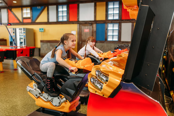 Дві маленькі дівчата грають в ігровий автомат, дитячий розважальний центр. Захоплені діти розважаються на дитячому майданчику в приміщенні. Діти грають на мотоциклах, розважальному центрі
 - Фото, зображення