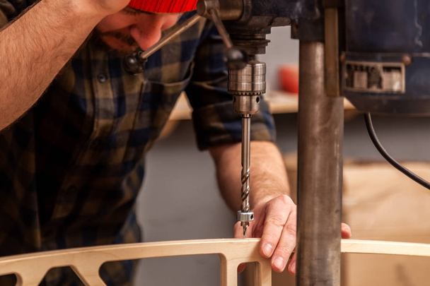 Primer plano de un hombre carpintero en un sombrero y una camisa está tallando una tabla de madera en una gran máquina de perforación en una vista lateral del taller, en el fondo una gran cantidad de herramientas
 - Foto, imagen