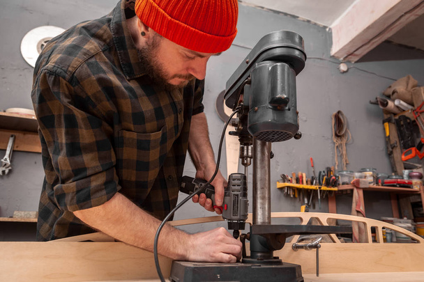 Close-up de carpinteiro experiente em roupas de trabalho e pequeno proprietário buiness está esculpir uma placa de madeira em uma broca de mão moderna em uma oficina de luz vista lateral, no fundo um monte de ferramentas
 - Foto, Imagem