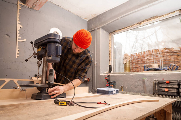 作業服、大工のキャップを持つ男は光のワーク ショップでモダンな大型掘削機に木の板を彫刻します。 - 写真・画像
