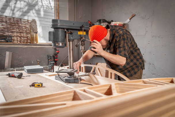 Um homem carpinteiro em um chapéu e uma camisa está esculpir uma placa de madeira em uma grande máquina de perfuração em uma oficina vista lateral, no fundo um monte de ferramentas
 - Foto, Imagem