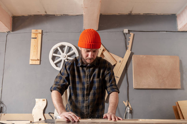 Молодой человек в оранжевой шляпе плотник в рабочей одежде обрабатывает деревянную доску с фрезерным станком в мастерской, вокруг много оборудования, деревянные доски
 - Фото, изображение