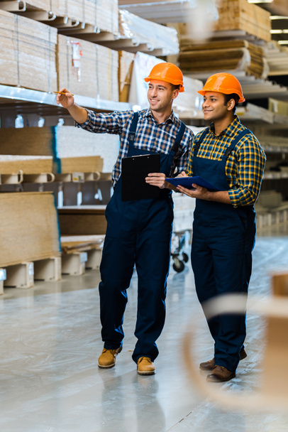 smiling warehouse worker in uniform pointing with finger near indian colleague - Photo, Image