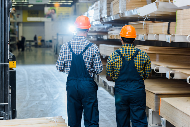 back view of two multicultural workers in overalls, plaid shirts and helmets in warehouse - Foto, Imagem