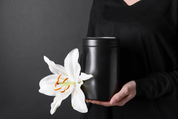 Woman with mortuary urn and lily flower on dark background, closeup - Photo, Image