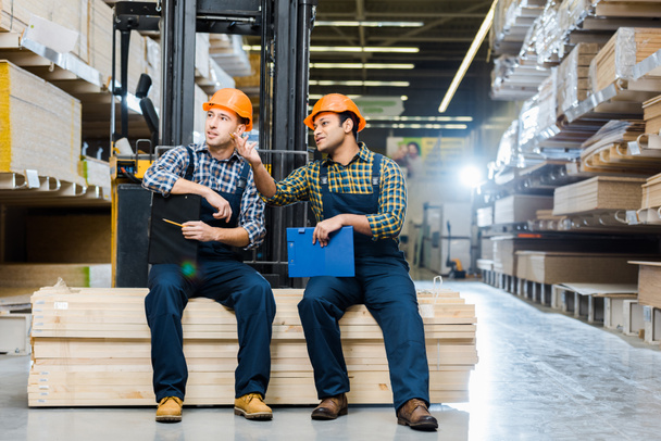 two multicultural workers talking while sitting on plywood in warehouse - Photo, Image