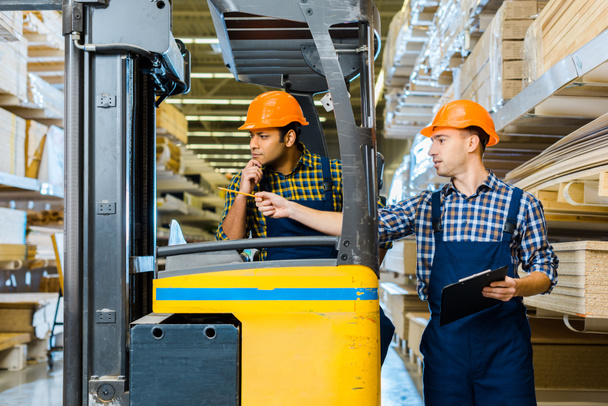 indian warehouse worker sitting in forklift machine near colleague pointing with pencil - Photo, Image