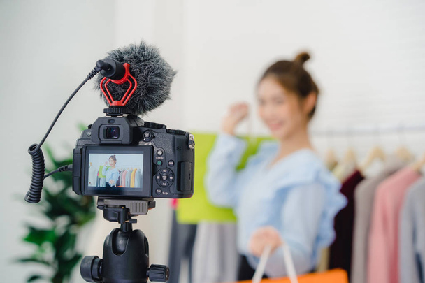 Asian fashion female blogger online influencer holding shopping bags and lots of clothes on clothes rack for recording new fashion video broadcast live video to social network by internet at home. - Valokuva, kuva