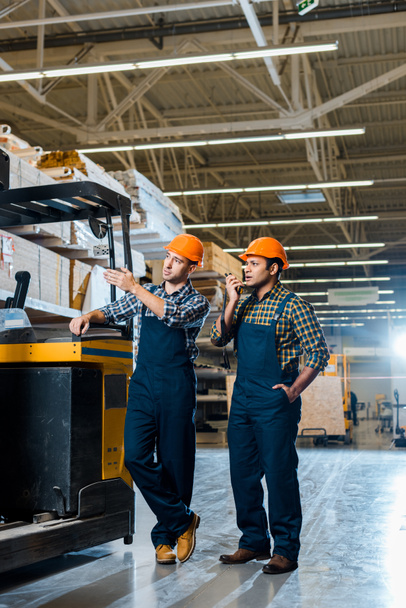 concentrated multicultural workers in helmets and plaid shirts standing near forklift machine - Photo, Image