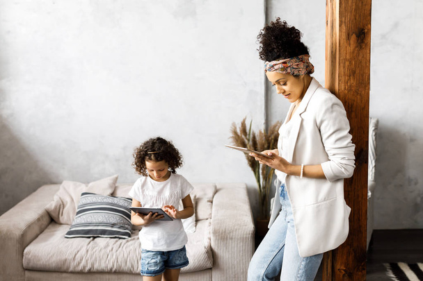A cute mother and daughter talk and look at the tablet while standing in the living room - Фото, изображение