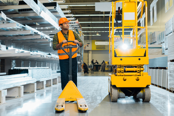 serious indian worker in safety vest and helmet standing near pallet jack in warehouse - Photo, Image